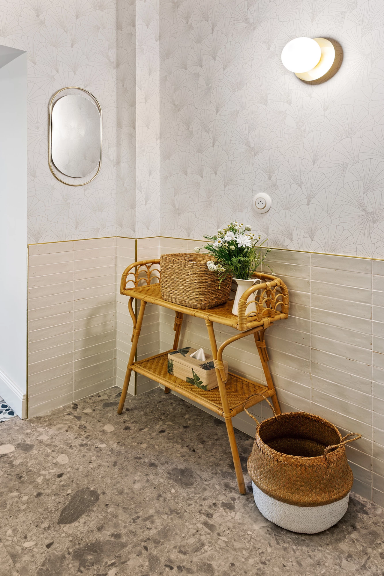 Coin toilettes lumineux avec papier peint clair à motif éventail, soubassement carrelé beige et console en rotin avec panier.