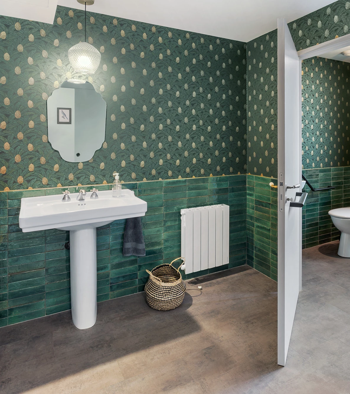 Salle de bain verte avec papier peint à motifs ananas, faïence verte, lavabo sur pied blanc et miroir vintage.