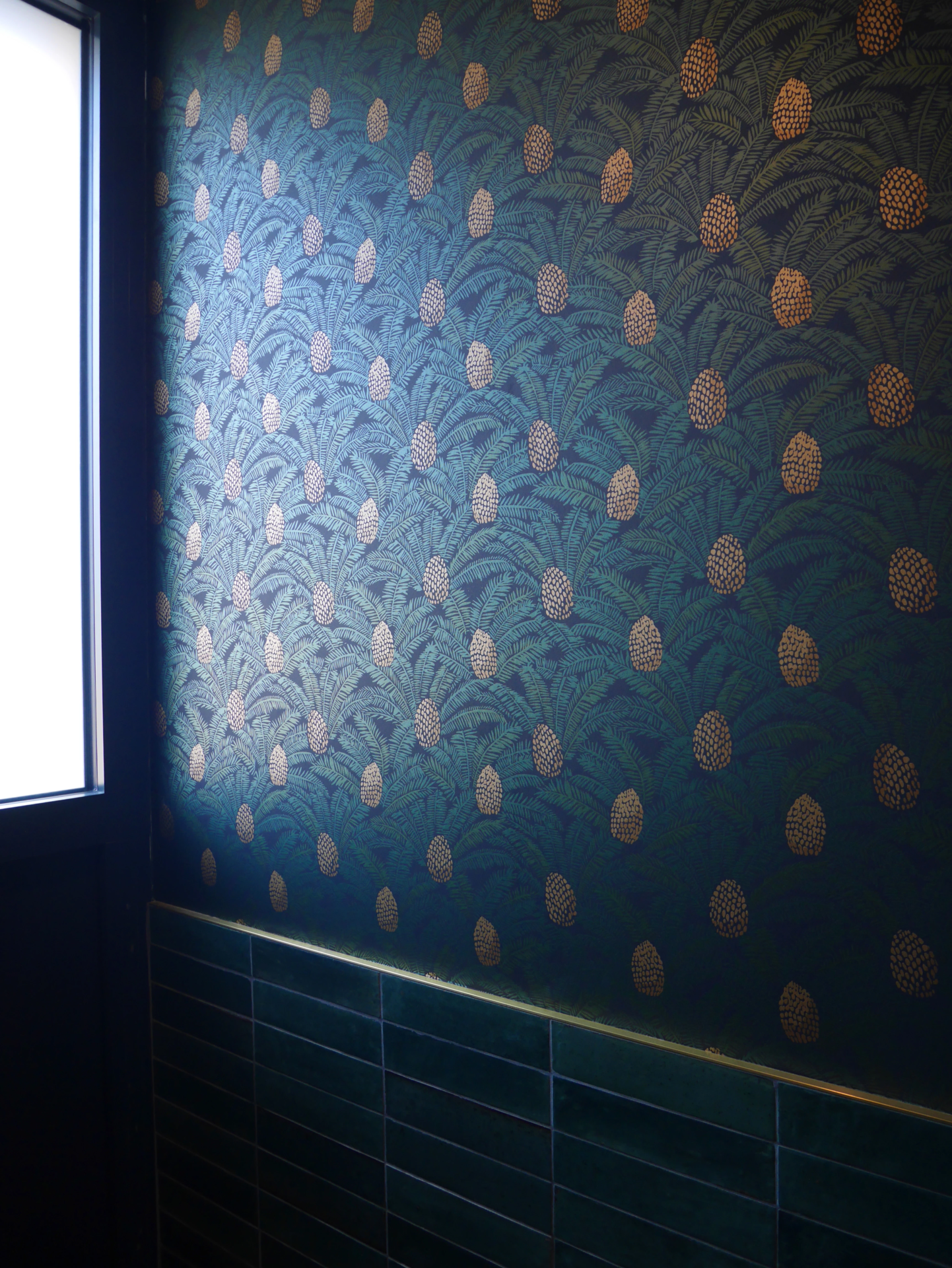 Détail de mur de salle de bain avec papier peint vert à motifs ananas et faïence verte éclairé par une fenêtre.