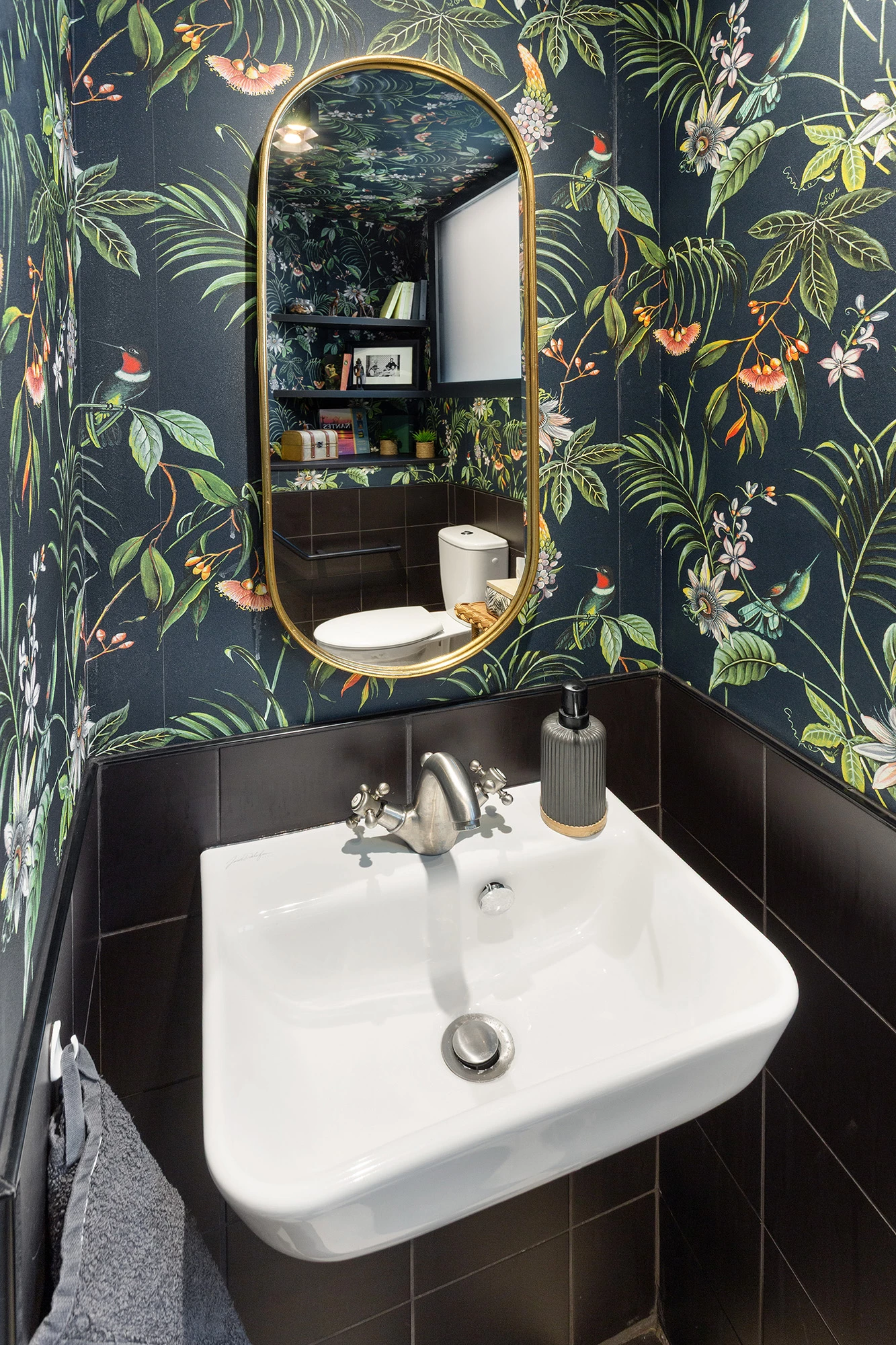 Petit lavabo blanc mural avec robinet rétro sous miroir ovale doré, papier peint tropical sombre et faïence noire.
