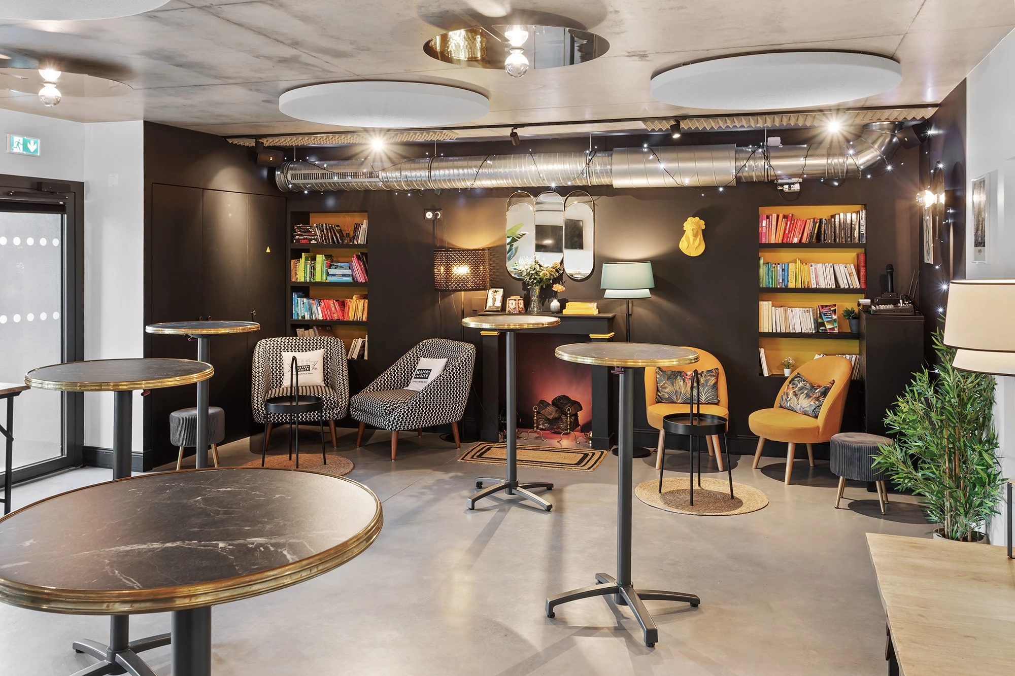 Espace lounge contemporain avec murs noirs, bibliothèque intégrée, fauteuils cosy et tables rondes effet marbre.