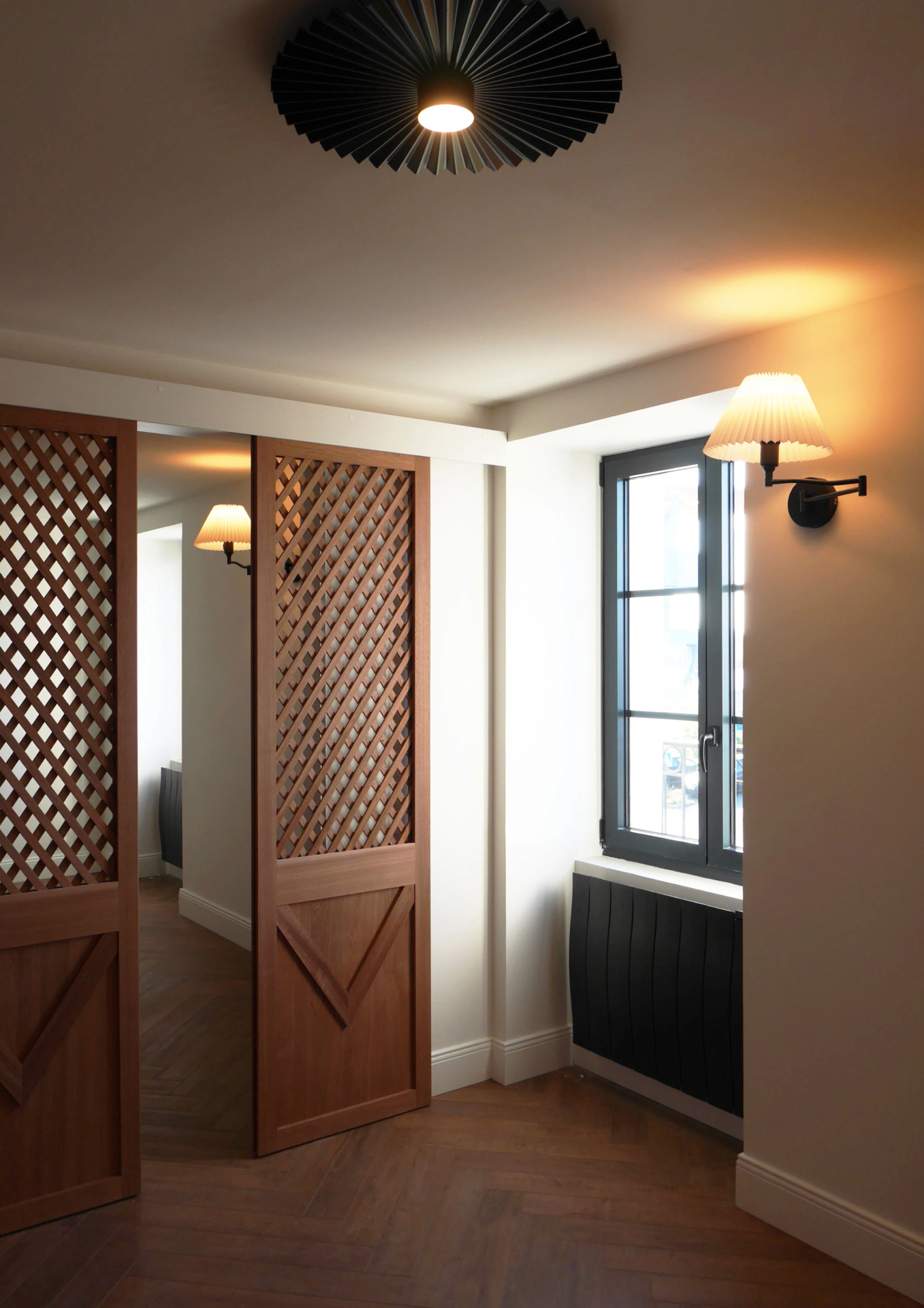 Paroi coulissante en bois à motif treillage séparant un espace intérieur, avec éclairage mural et fenêtre.
