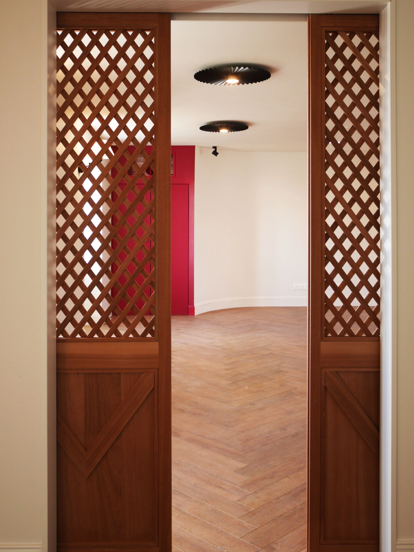Vue frontale de portes coulissantes en bois ajouré type treillage ouvrant sur une pièce au parquet en chevrons.